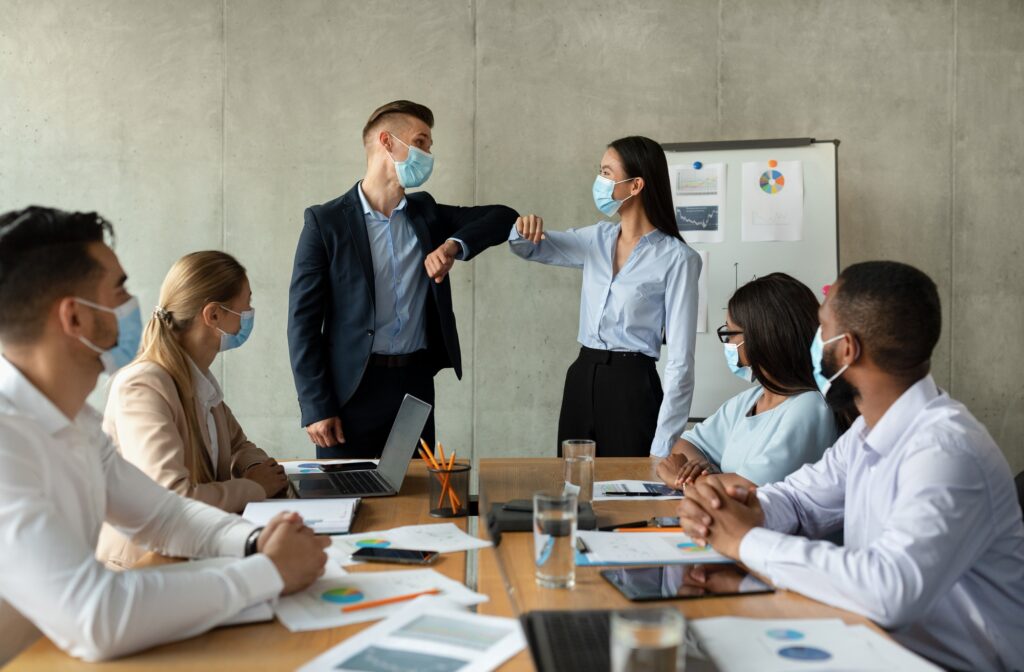 Business Communication During Pandemic. Coworkers In Medical Masks Bumping Elbows In Office